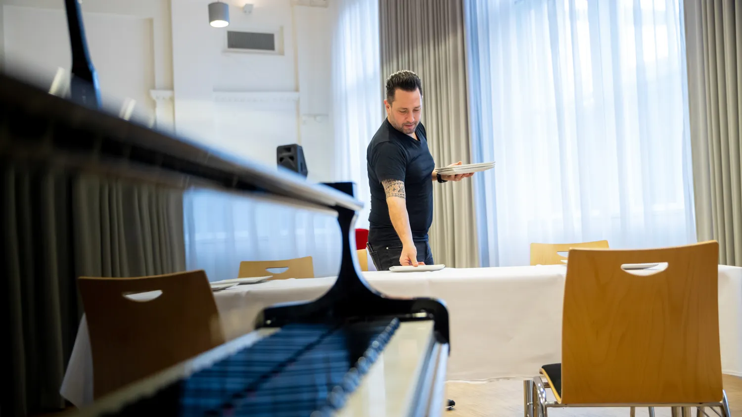 Man in zwart shirt dekt tafel. Piano vleugel op voorgrond