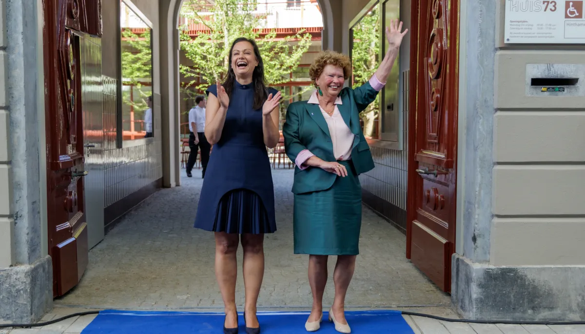 Twee vrouwen, wethouder en directeur, zwaaien uitnodigend vanaf blauwe loper