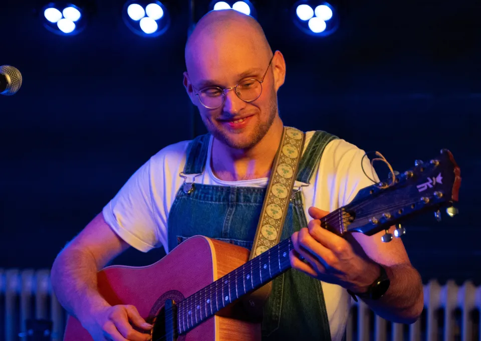 man kaal met gitaar op podium met lichten erboven lachend bril 