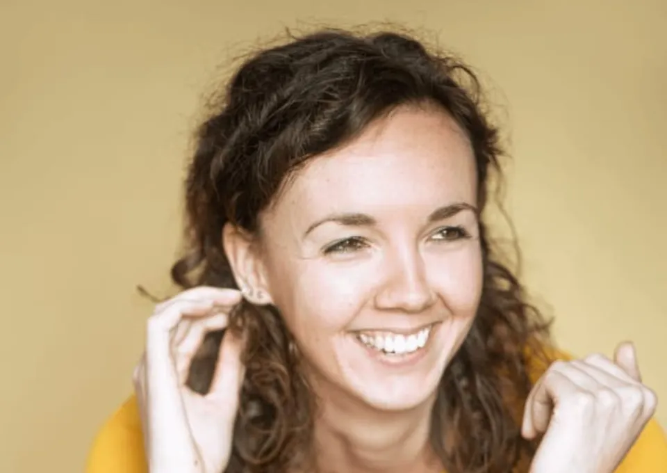 vrouw met krullend bruin haar lachend met hand bij oor geel shirt en gitaat op schoot 