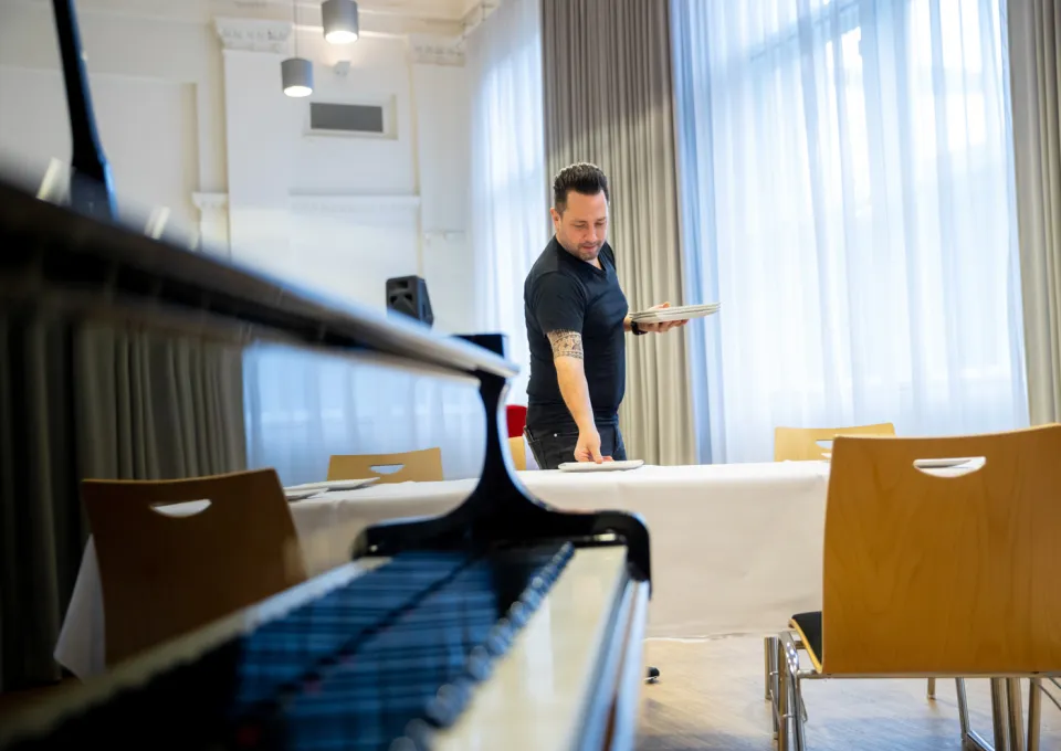 Man in zwart shirt dekt tafel. Piano vleugel op voorgrond