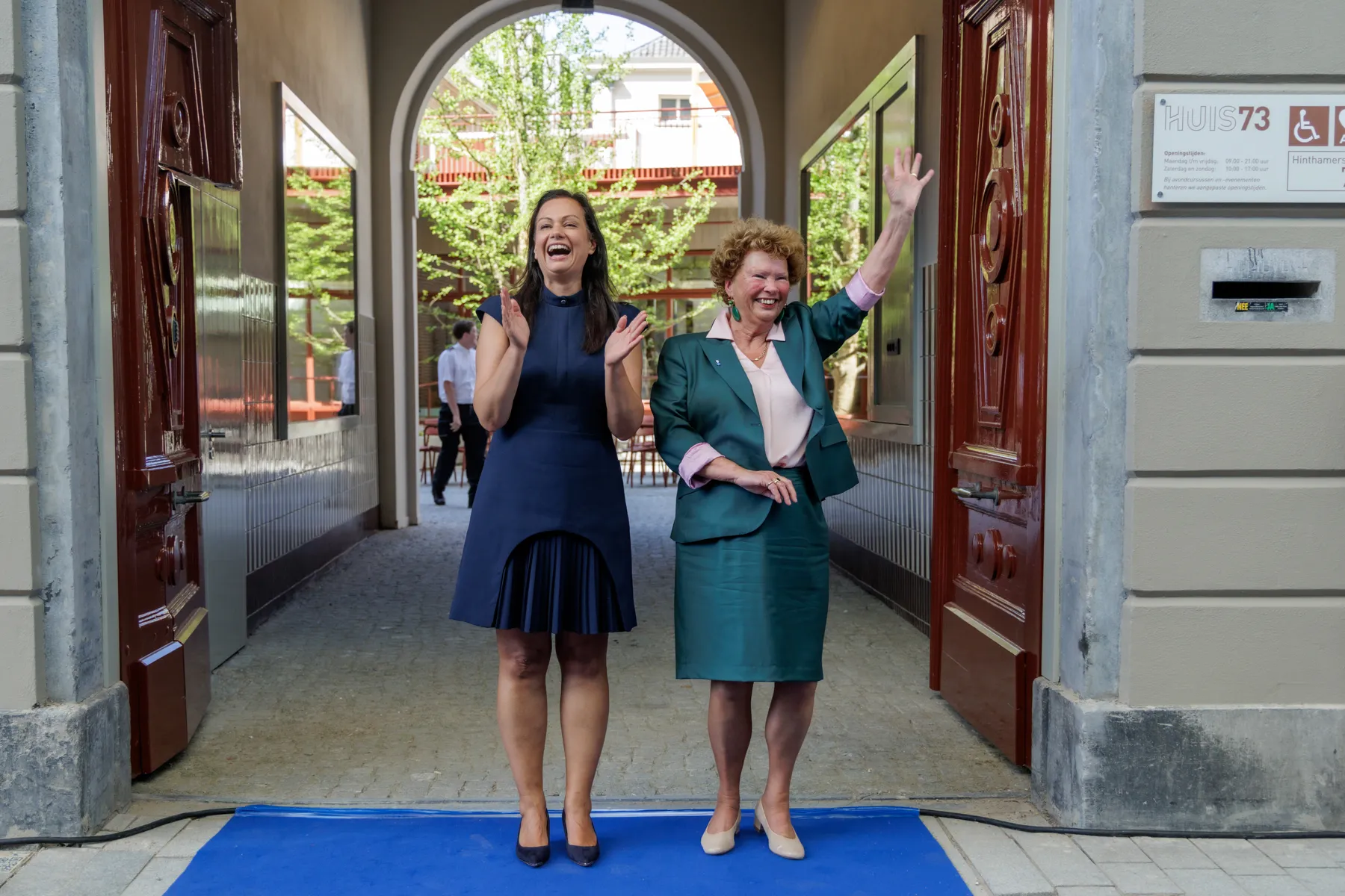 Twee vrouwen, wethouder en directeur, zwaaien uitnodigend vanaf blauwe loper