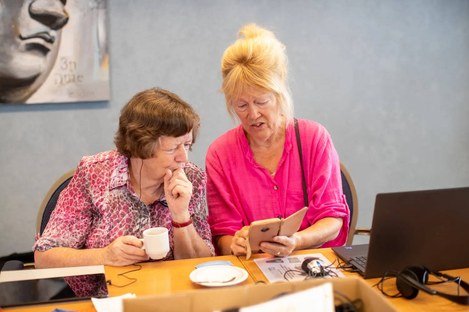 Twee deelnemers zitten aan een tafel tijdens een digitale workshop. Samen bekijken ze een smartphone. Op tafel staan een laptop, een kop koffie en cursusmateriaal.
