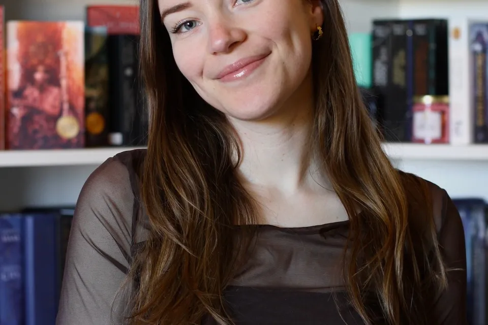 Portret Britt - jonge vrouw met lang bruin haar dat over de schouders hangt. Ze draagt zwarte kleding. Achter haar een boekenkast. 