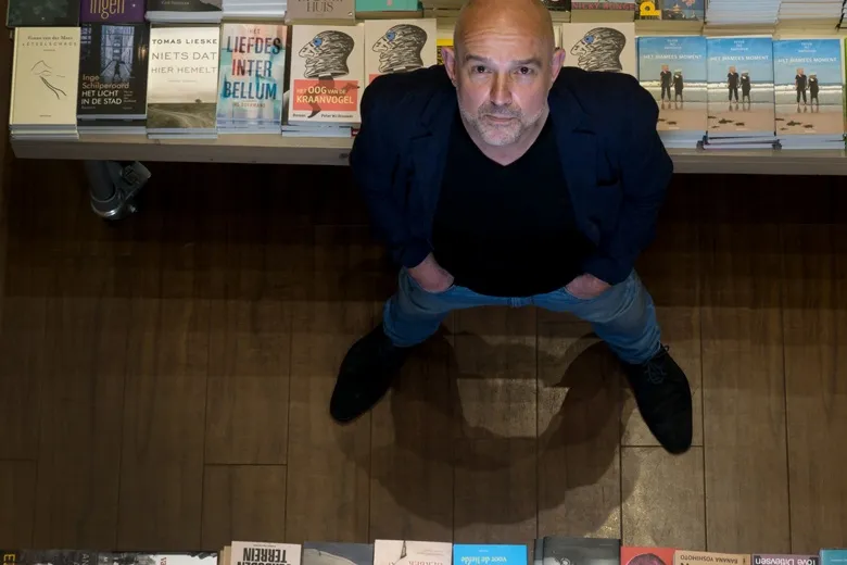 Kale man met grijze baard en zwarte blader van bovenaf gefotografeerd naast een stapel boeken