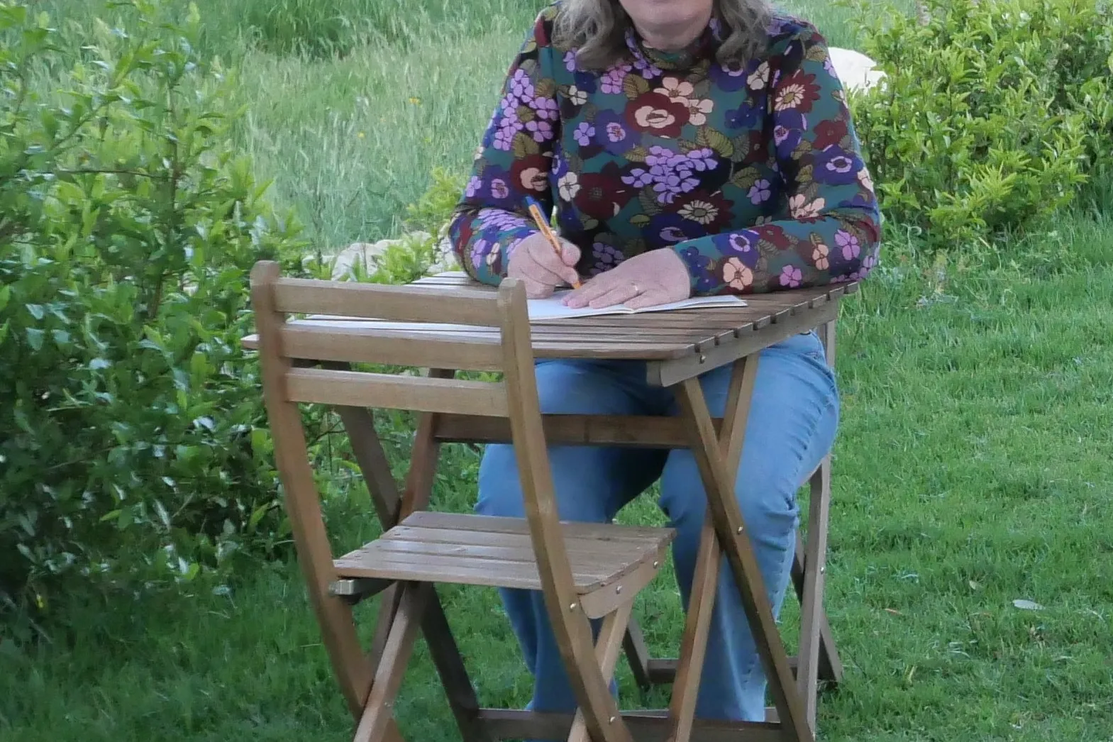 Vrouw zit aan een houten tafeltje te schrijven in de buitenlucht