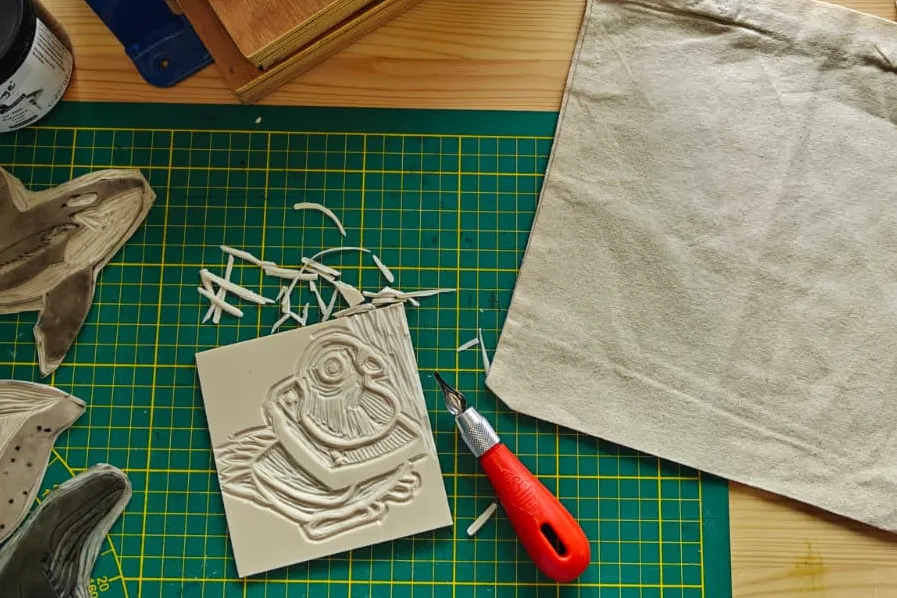 Een linoafdruk, met daarbij een canvas tasje en een drukpers. 