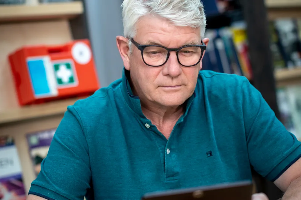 Man met grijs haar, bril en blauwe polo kijkt naar zijn tablet