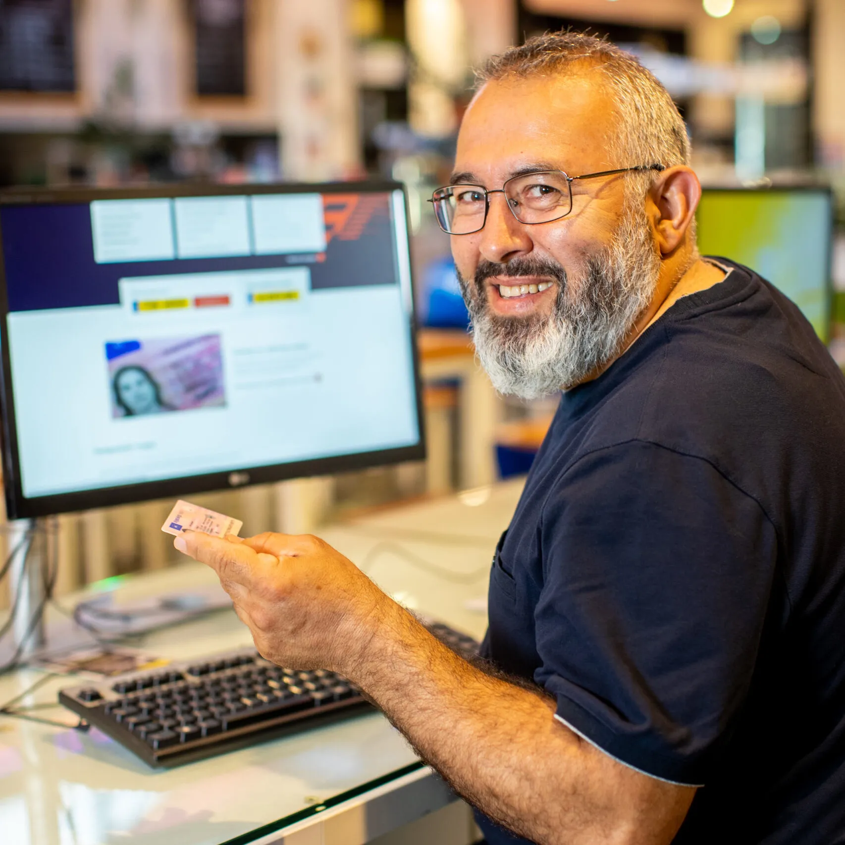 Man van ongeveer 50 jaar oud zit achter computer en heeft zijn rijbewijs in zijn linkerhand. De man kijkt lachend achterom de camera in.