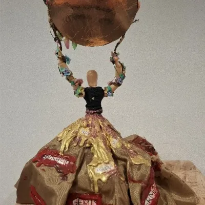 Kunstwerk van houten poppetje met stoffen jurk en koperkleurige cirkel