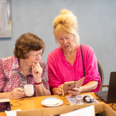 Twee deelnemers zitten aan een tafel tijdens een digitale workshop. Samen bekijken ze een smartphone. Op tafel staan een laptop, een kop koffie en cursusmateriaal.