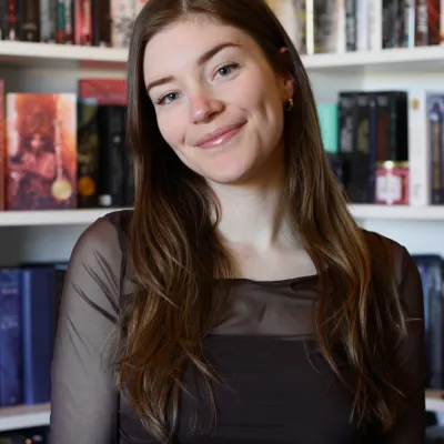 Portret Britt - jonge vrouw met lang bruin haar dat over de schouders hangt. Ze draagt zwarte kleding. Achter haar een boekenkast. 