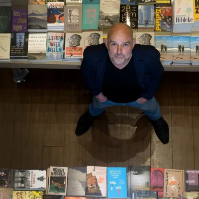 Kale man met grijze baard en zwarte blader van bovenaf gefotografeerd naast een stapel boeken