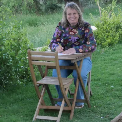 Vrouw zit aan een houten tafeltje te schrijven in de buitenlucht